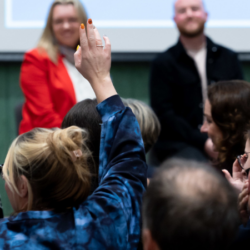 A photo of a woman raising her hand in a seated audience
